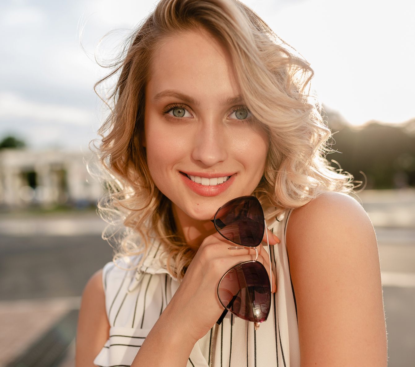 Smiling woman holding sunglasses outdoors, sunny day.